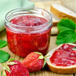 strawberry jam in a jar and on some toast. There's a knife on the toast after spreading it and a couple of strawberries on the counter.