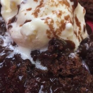 A close-up of a decadent Hot Fudge Chocolate Pudding Cake with a scoop of vanilla ice cream on top, drizzled with chocolate sauce and served over rich, gooey chocolate cake on a red plate.
