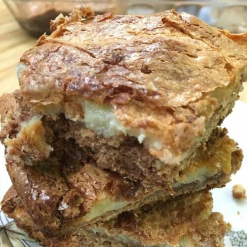 Three gooey Spice Cake Pecan Chess Squares with a golden, cracked crust are stacked on a plate, revealing a dense, moist layer beneath the flaky top. In the background, more tempting bars wait in the baking dish.