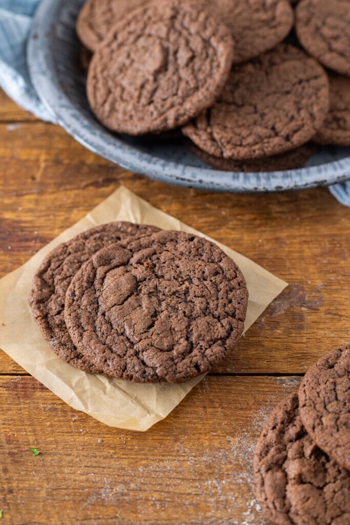 Three chocolate cookies on parchment paper rest on a wooden surface, with more cookies piled in a blue-grey ceramic plate in the background, all made from an easy, no chill chewy chocolate cookie recipe.