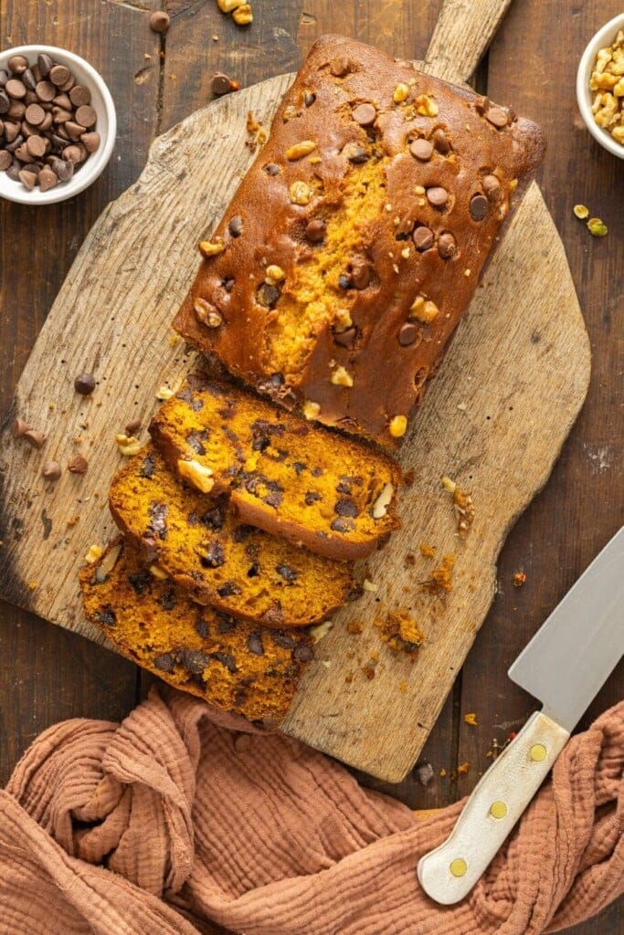 A loaf of pumpkin bread with chocolate chips and nuts sits on a wooden board, partially sliced—one of our 7 Easy Pumpkin Recipes. A knife rests nearby, with bowls of chocolate chips and nuts and a rust-colored cloth beside the board.