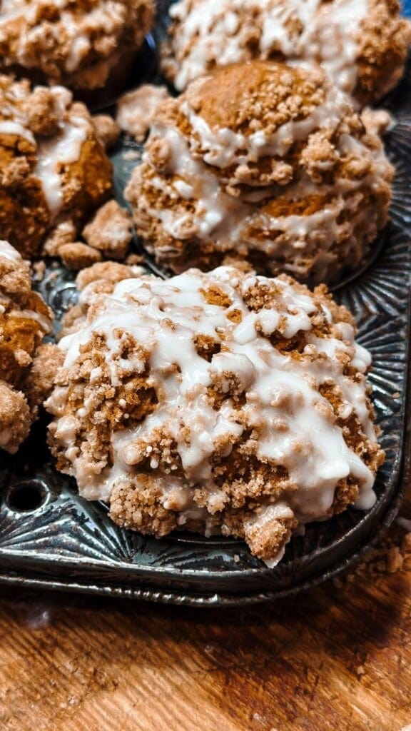Close-up of freshly baked coffee cake muffins with crumbly streusel topping and a generous drizzle of white glaze, perfect inspiration if you love 7 Easy Pumpkin Recipes, sitting in a dark metal muffin tin on a wooden surface.