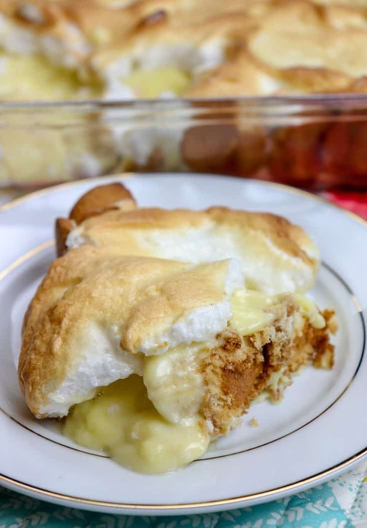 A slice of homemade banana pudding topped with toasted meringue on a white plate, with a larger glass baking dish of the dessert in the background.