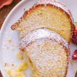 Two slices of Old Fashioned Cream Cheese Pound Cake dusted with powdered sugar rest on a white plate, served with fresh blackberries and a strawberry. A forkful of cake sits nearby, ready to enjoy.