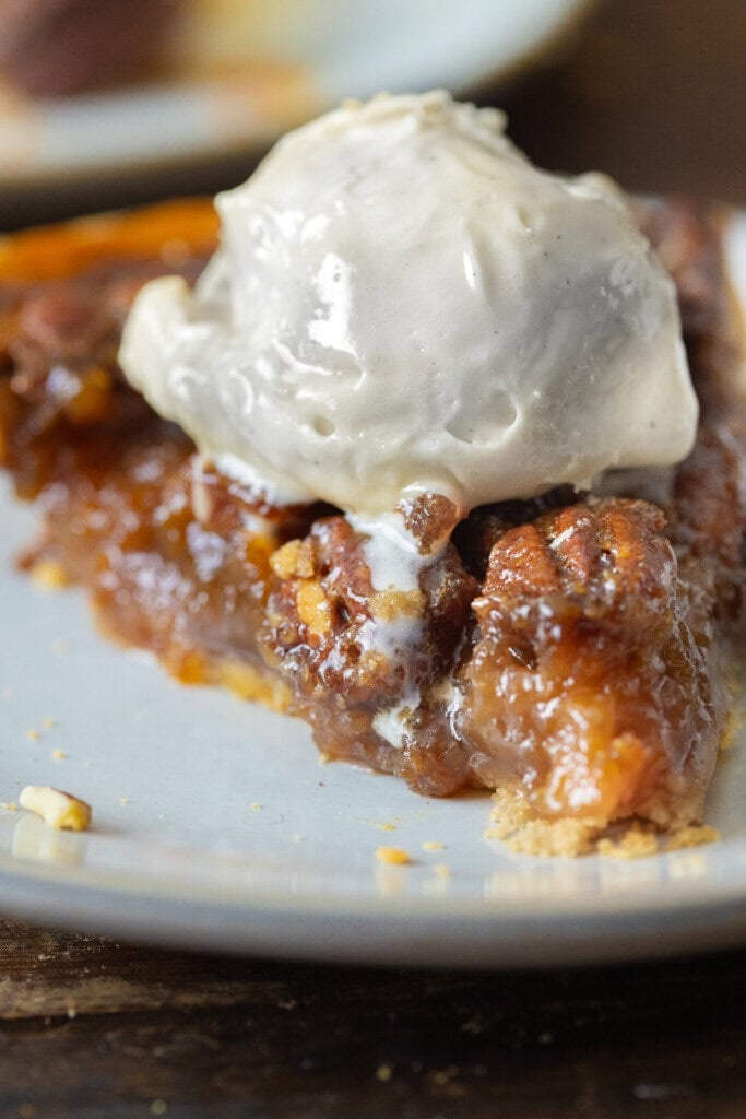 A close-up of a slice from an Old Fashioned Pecan Pie Recipe topped with a scoop of vanilla ice cream. The filling appears gooey and rich, with visible pecans and a perfectly flaky crust on a white plate.