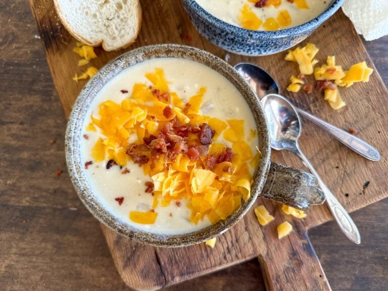 A bowl of creamy easy slow cooker potato soup topped with shredded cheddar cheese and crispy bacon bits, set on a wooden board with two spoons, a slice of bread, and another bowl of soup in the background.