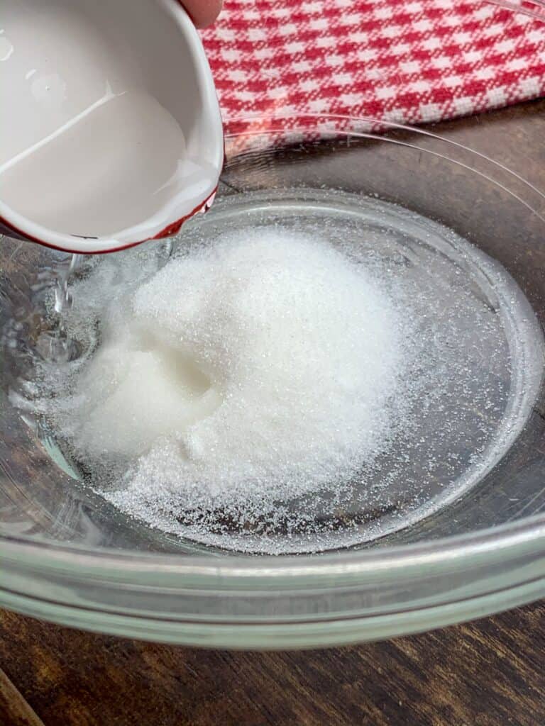 A clear glass bowl containing white granulated sugar, a key ingredient in this Easy Copycat Chick-Fil-A Coleslaw Recipe (Authentic), with white vinegar being poured onto it. A red-and-white checkered cloth sits on the wooden surface in the background.