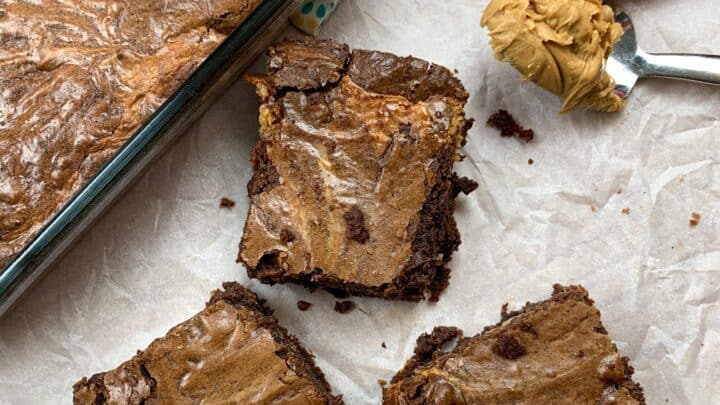 Three brownies sitting next to a pan of brownies and a spoonful of peanut butter