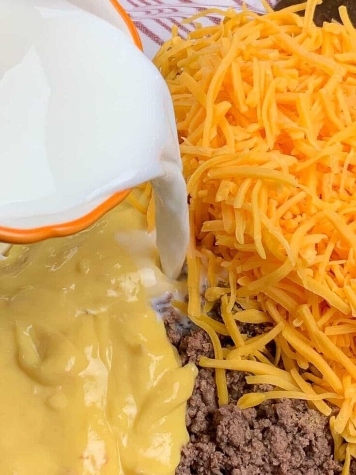 A close-up of shredded cheddar cheese, cooked ground beef, and condensed soup in a bowl, with milk being poured over the ingredients for The Best Tater tot casserole recipe.