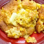A serving of golden brown Jiffy Corn Pudding with visible corn kernels and a pat of melting butter on top, presented on a red plate.