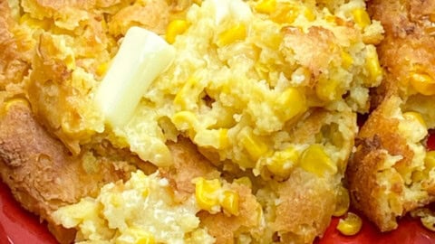 A serving of golden brown Jiffy Corn Pudding with visible corn kernels and a pat of melting butter on top, presented on a red plate.