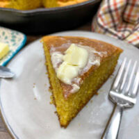 Cornbread with butter on top. The cornbread has some butter melting on the top and there is a fork next to the cornbread. The cornbread is sitting on a white plate.