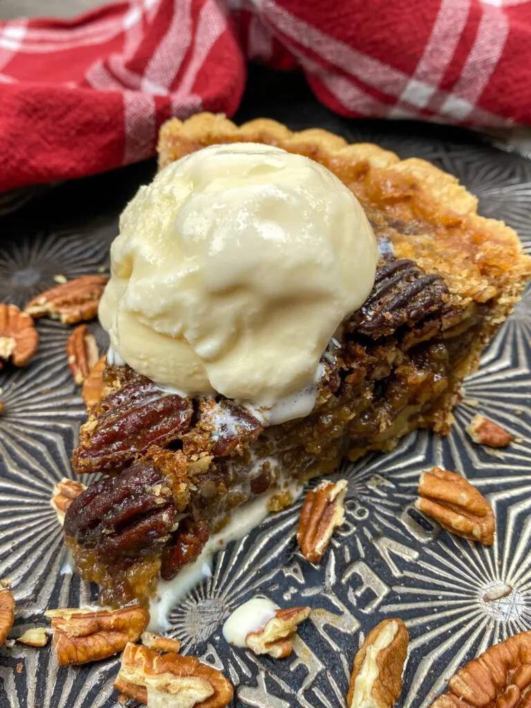 A slice of pecan pie without corn syrup on a cookie sheet. The pie has a scoop of vanilla ice cream on top and there is a red and white towel in the background.