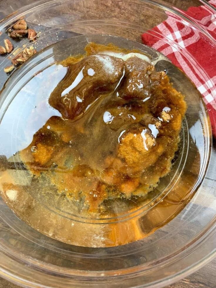 A glass bowl containing a Karo syrup and brown sugar, sits on a wooden surface next to a red and white checkered clothβperfect for making an Easy Pecan Pie Cobbler Recipe. Some chopped pecans are visible in the corner.
