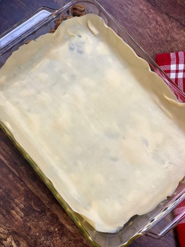 A rectangular glass baking dish filled with an Easy Pecan Pie Cobbler Recipe, covered with an unbaked sheet of pastry dough. The dish sits on a wooden surface next to a red and white checkered cloth.