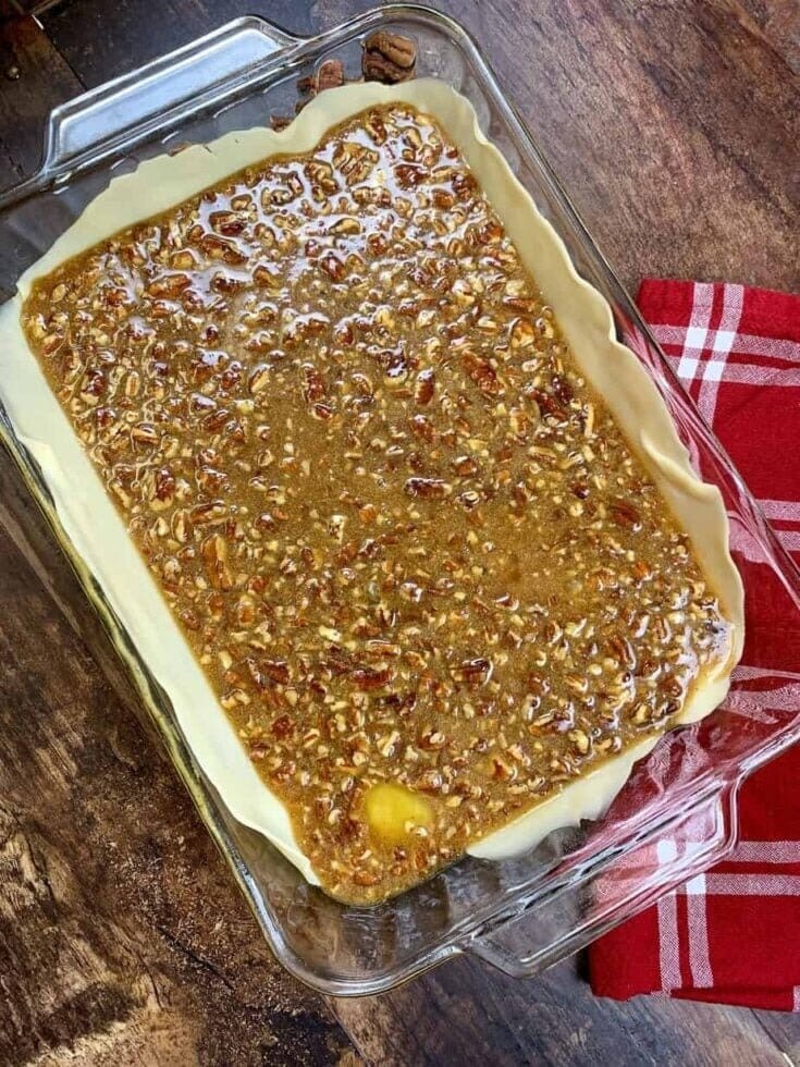 A glass baking dish lined with dough and filled with an Easy Pecan Pie Cobbler Recipe mixture sits on a wooden surface next to a folded red and white checkered kitchen towel.