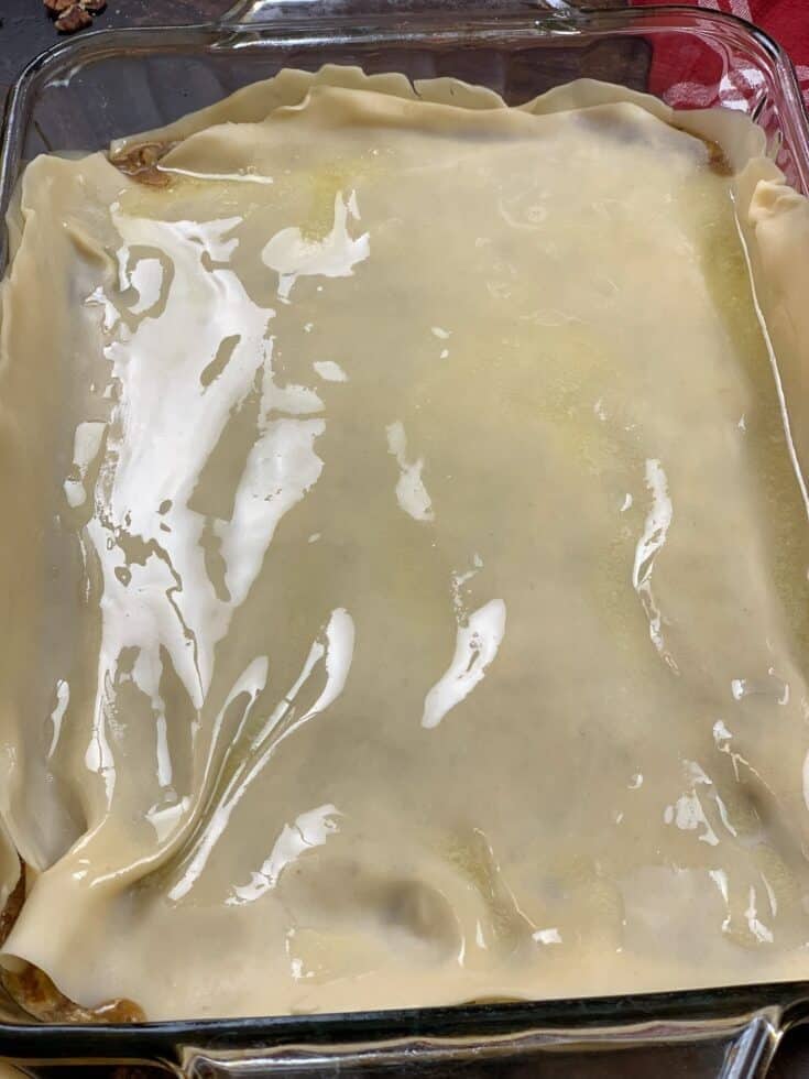 A glass baking dish filled with an Easy Pecan Pie Cobbler Recipe, topped with an uncooked sheet of pastry dough that is slightly shiny and draped over the edges.