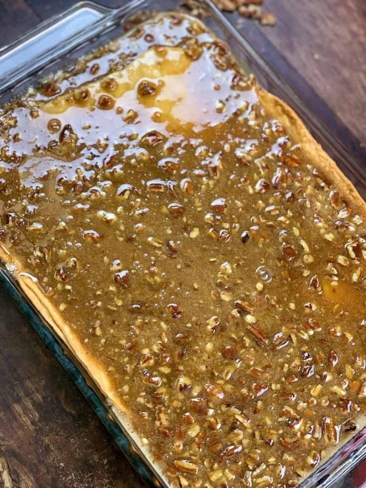 A close-up of a rectangular glass baking dish filled with a gooey Easy Pecan Pie Cobbler Recipe. The surface is glossy, caramelized brown, and sprinkled with chopped pecans. The dish rests on a dark wooden surface.