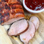 pork loin sliced on a wooden cutting board with some barbecue sauce in a small bowl next to it.