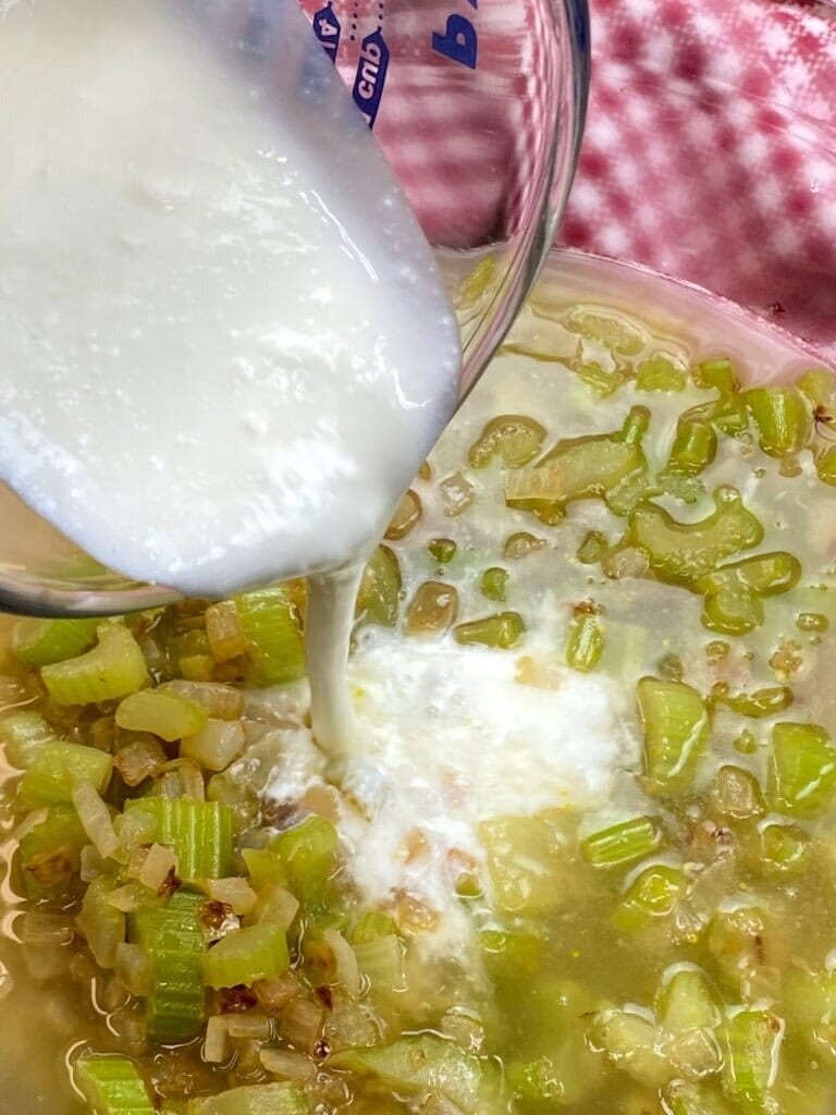 A glass measuring cup pours buttermilk into a pot of chopped celery, onions, and broth—the perfect base for a classic Jiffy Cornbread Dressing Recipe—set against a red and white checkered cloth.