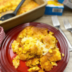 A serving of Jiffy corn pudding with a golden crust is plated on a red dish beside a baking pan, with forks and a napkin nearby. Text at the top reads “Easy Jiffy Corn Pudding Recipe.”.