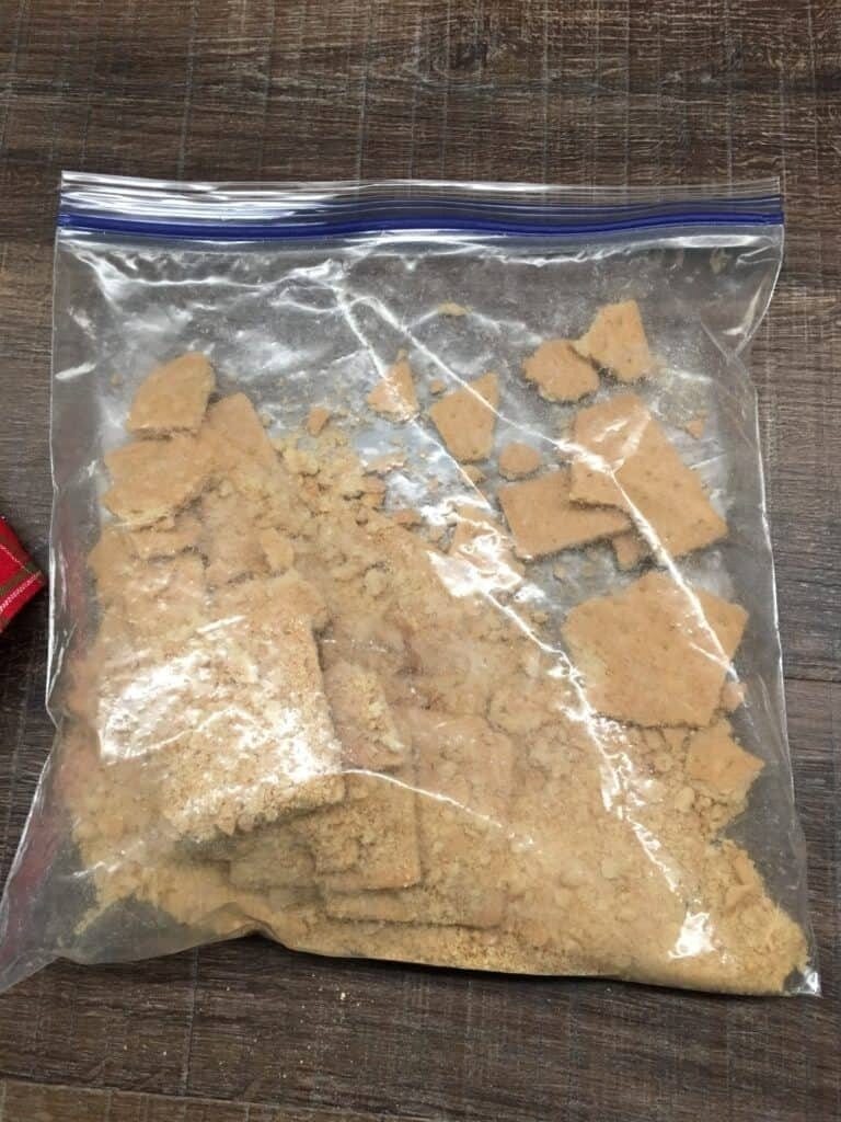 A clear resealable plastic bag contains broken pieces and crumbs of graham crackers, sitting on a wooden surface—perfect for learning how to make hello dolly bars.