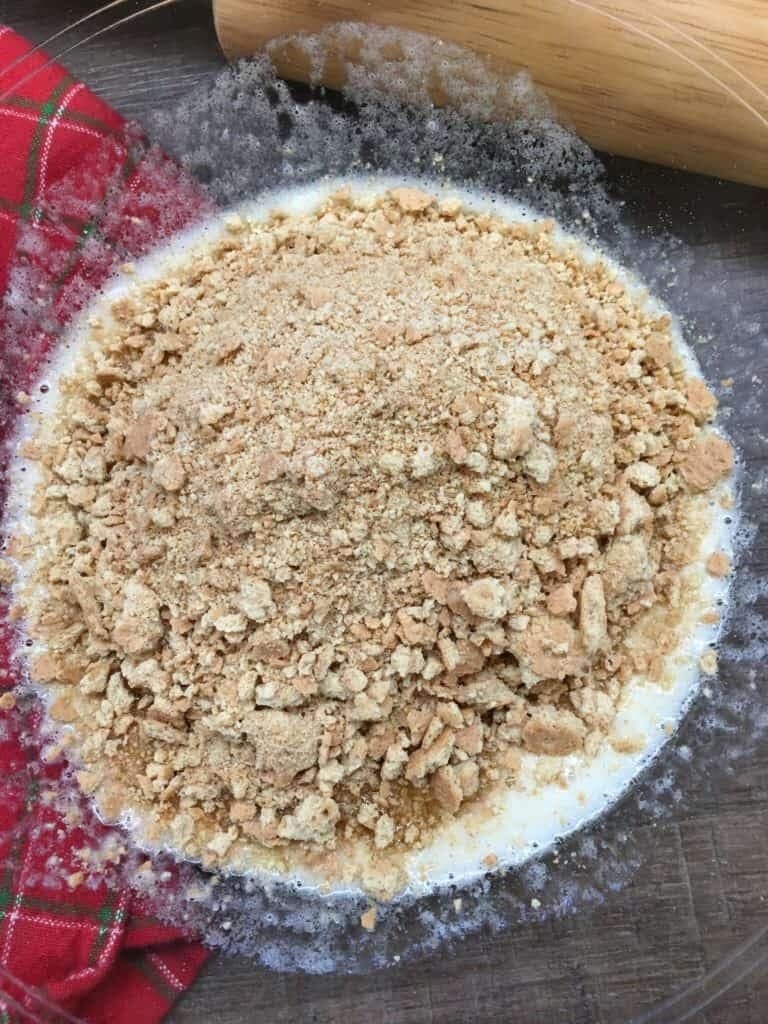 A glass bowl filled with crushed graham crackers atop a creamy mixture, showing the first step of how to make hello dolly bars, sits next to a red plaid cloth and wooden rolling pin on a dark countertop.