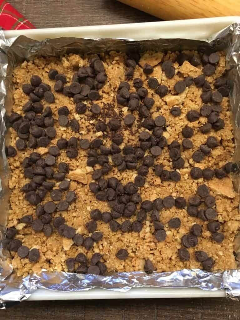 A square baking pan lined with foil contains a crumbly dessert topped with scattered chocolate chips—classic for those learning how to make hello dolly bars. The pan rests on a wooden surface, with a rolling pin partially visible at the top.