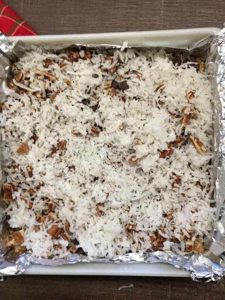 A baking dish lined with foil, filled with a dessert ready to be baked—this is how to make hello dolly bars, topped with shredded coconut, chopped nuts, and chocolate chips.