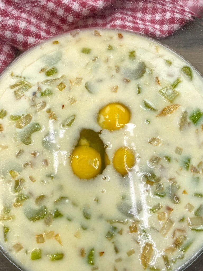 A close-up of a bowl containing a mixture of chopped green vegetables and onions in a creamy liquid, with three raw eggs cracked on top—perfect prep for a classic Jiffy Cornbread Dressing Recipe—set against a red and white checkered cloth.