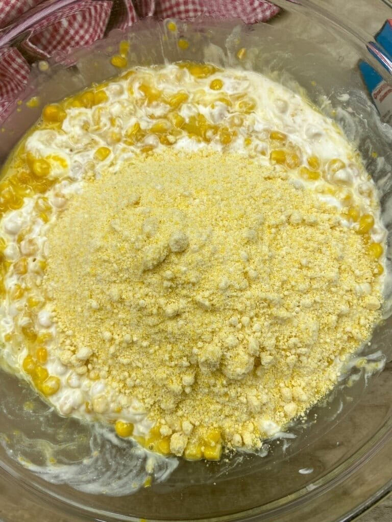 A glass bowl contains a mixture of corn kernels, creamy ingredients, and a mound of cornmeal, ready to be mixed for Jiffy Corn Pudding. A red checked cloth is partially visible in the background.