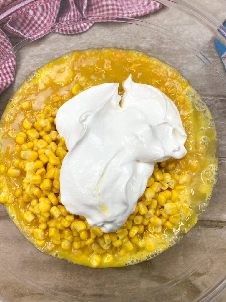A clear bowl with yellow corn kernels, beaten eggs, and a large dollop of white sour cream sits on a light wooden surface with a red-and-white checkered clothโan inviting start for Jiffy Corn Pudding.