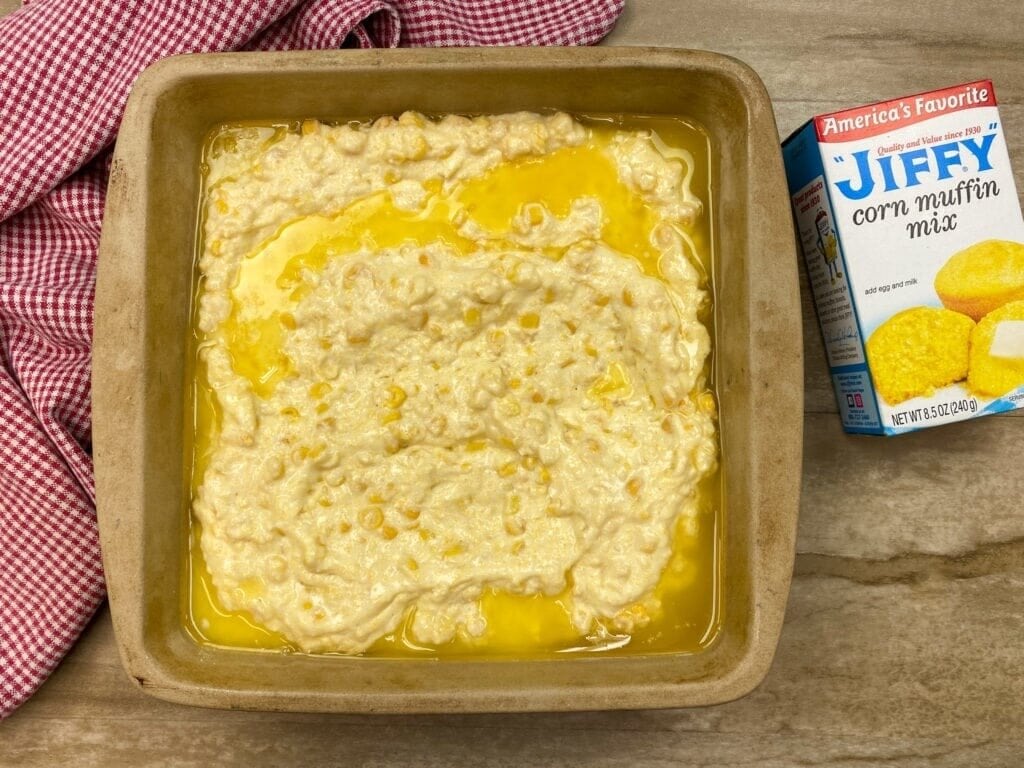 A square baking dish filled with Jiffy Corn Pudding batter topped with melted butter sits on a wooden surface. A red and white checkered cloth and a box of Jiffy corn muffin mix are beside the dish.