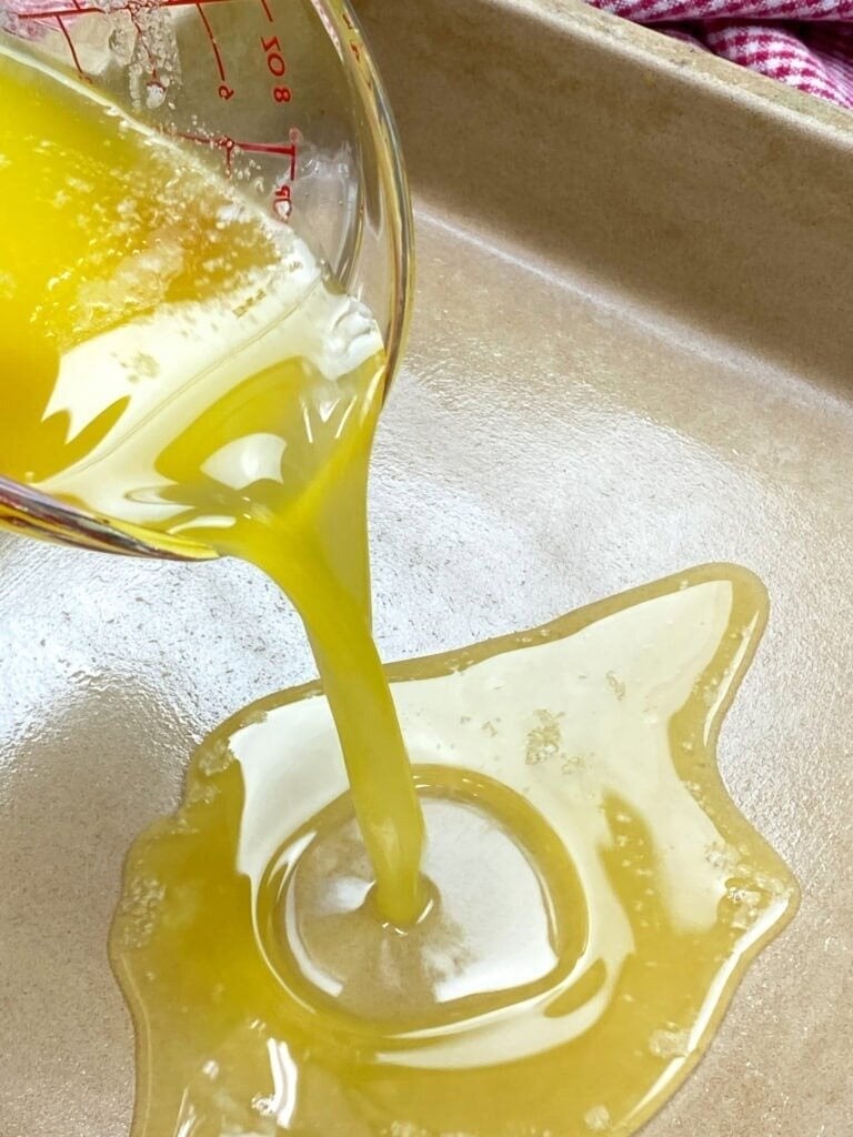 Melted butter is being poured from a glass measuring cup into a beige baking dish, spreading across the bottom in a golden poolโthe first step for making delicious Jiffy Corn Pudding.