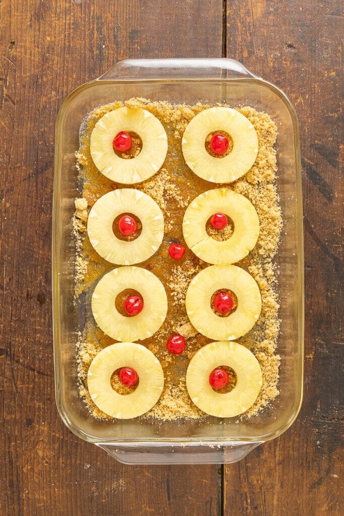 A glass baking dish on a wooden surface holds a layer of brown sugar, pineapple rings, and maraschino cherries—perfect for creating a classic Pineapple Upside Down Cake Recipe With Cake Mix.