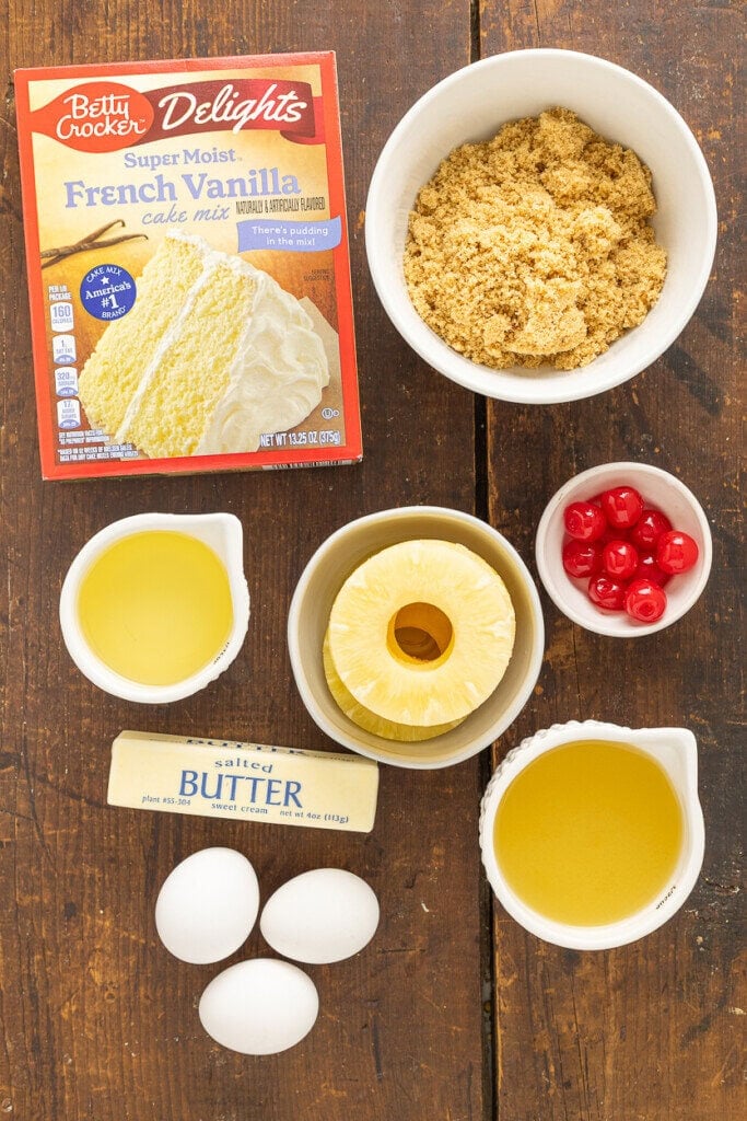 A box of French vanilla cake mix, brown sugar, maraschino cherries, pineapple rings and juice, butter, eggs, and vegetable oil—everything you need for a delicious Pineapple Upside Down Cake Recipe With Cake Mix on a wooden table.