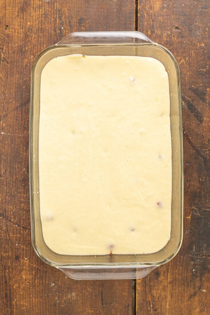 A glass baking dish filled with smooth, pale yellow Pineapple Upside Down Cake Recipe With Cake Mix batter sits on a rustic wooden surface, ready to be baked.