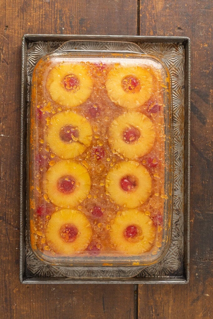 A rectangular glass baking dish holds a Pineapple Upside Down Cake Recipe With Cake Mix, topped with pineapple rings and red cherries, set on a vintage metal tray on a rustic wooden table.