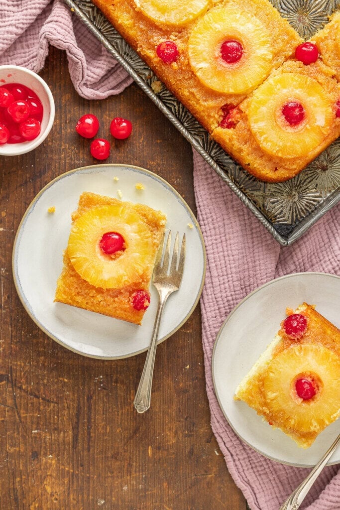 Two slices of pineapple upside-down cake with cherries on top sit on plates with forks. The rest of this Pineapple Upside Down Cake Recipe With Cake Mix is on a tray, with a bowl of maraschino cherries and a pink cloth on the wooden table.