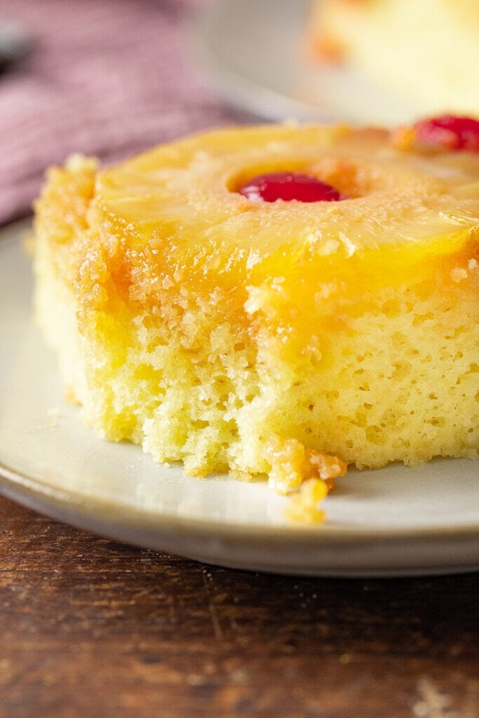 A close-up of a slice of Pineapple Upside Down Cake made from a cake mix, topped with a pineapple ring and red cherry, served on a light-colored plate.