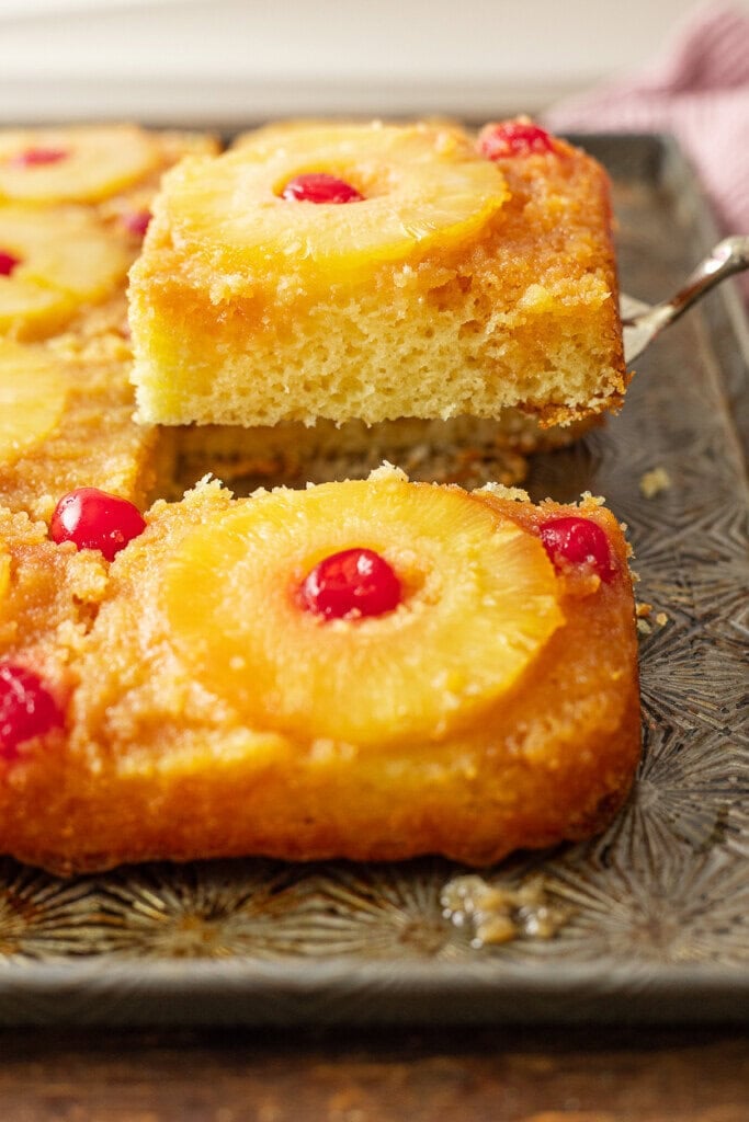 A close-up of a Pineapple Upside Down Cake Recipe With Cake Mix, topped with pineapple rings and cherries. A square slice is being lifted from the pan, revealing its moist, fluffy texture.