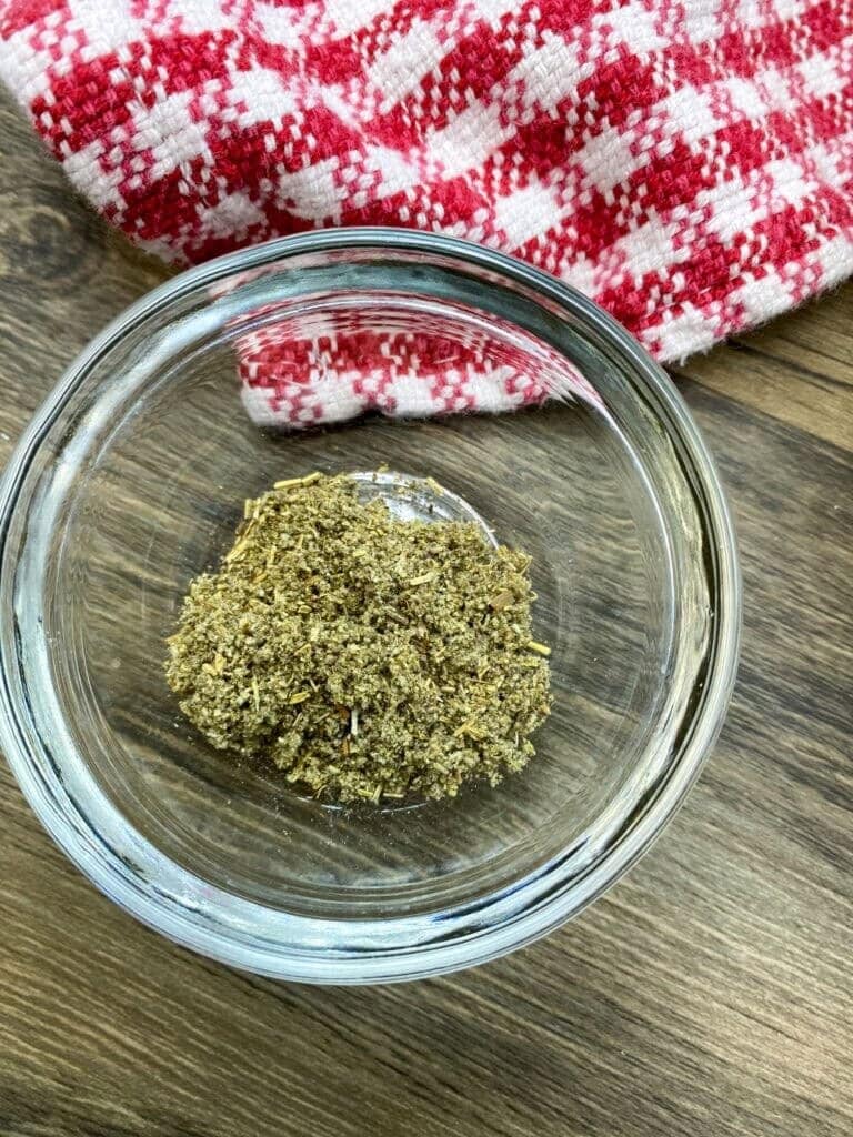 A small glass bowl containing a mixture of dried herbs for a Jiffy Cornbread Dressing Recipe sits on a wooden surface next to a red and white checkered cloth.