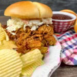 BBQ chicken on a bun with coleslaw. Potato chips are on the white plate next to the chicken.