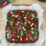 Chocolate fudge inside of a clear glass baking dish. The fudge is sitting inside of parchment paper. There are red, white, and green sprinkles on top.