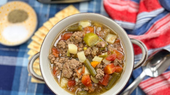 A bowl of hamburger vegetable soup recipe. There are some crackers on the side and a Crock Pot behind it.