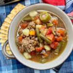 Hamburger soup in a bowl with veggies. There are some crackers on the side of the bowl.