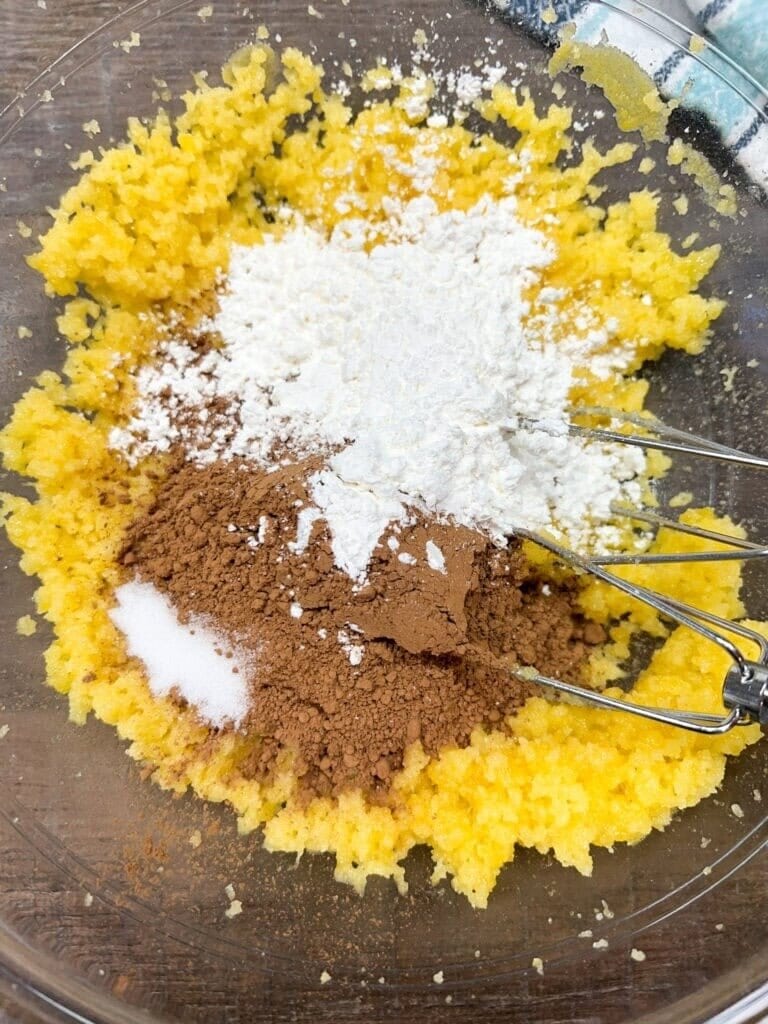 A glass bowl with yellow creamed mixture, topped with piles of flour, cocoa powder, and sugar—just the start of a delicious Homemade Chocolate Cream Pie Recipe. Metal beaters are partially visible, ready to mix the ingredients together.
