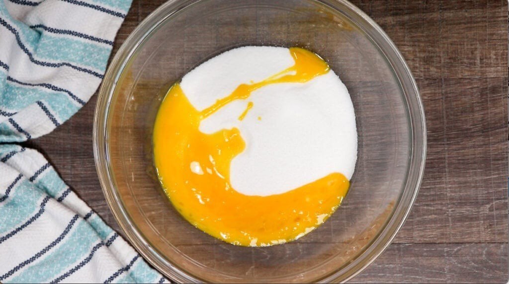 A clear glass bowl with granulated white sugar and partially mixed egg yolks sits on a wooden surface next to a blue and white striped kitchen towel—just the beginning of a delicious Homemade Chocolate Cream Pie Recipe.
