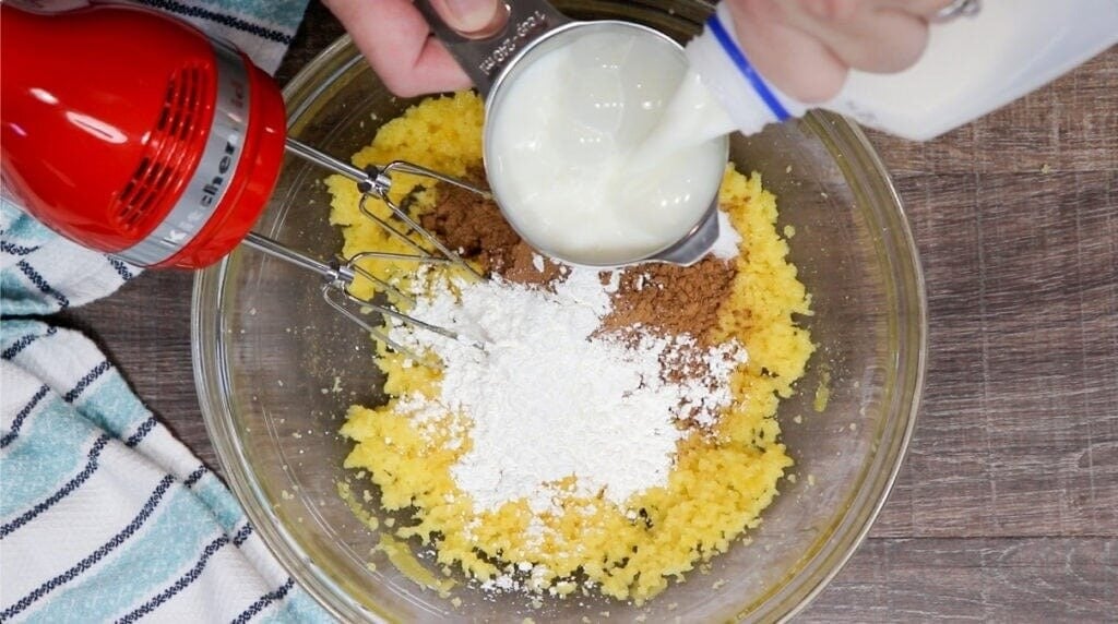 A hand pours milk from a bottle into a measuring cup over flour, cocoa powder, and yellow batter—perfect for a Homemade Chocolate Cream Pie Recipe. A red hand mixer rests in the bowl, with a striped towel nearby on the wooden surface.