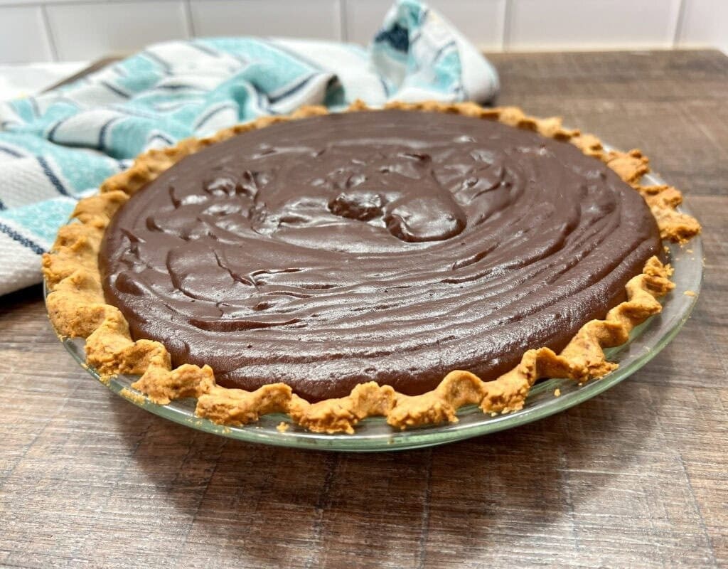 A glass pie dish filled with smooth, glossy chocolate pie from a homemade chocolate cream pie recipe is topped with a golden, crimped crust and sits on a wooden surface with a blue and white towel in the background.